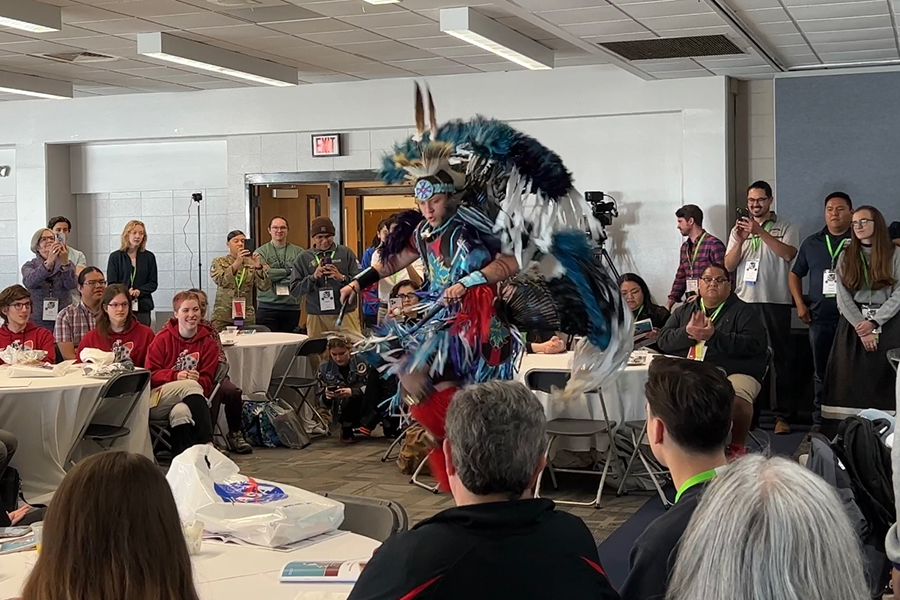 An Indigenous man wearing full Powwow regalia demonstrates a traditional dance.