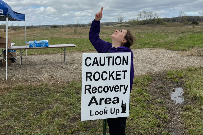 A&A student Grace Pardini points at the sky while standing behind a sign that reads, "Caution: Rocket recovery area. Look up"