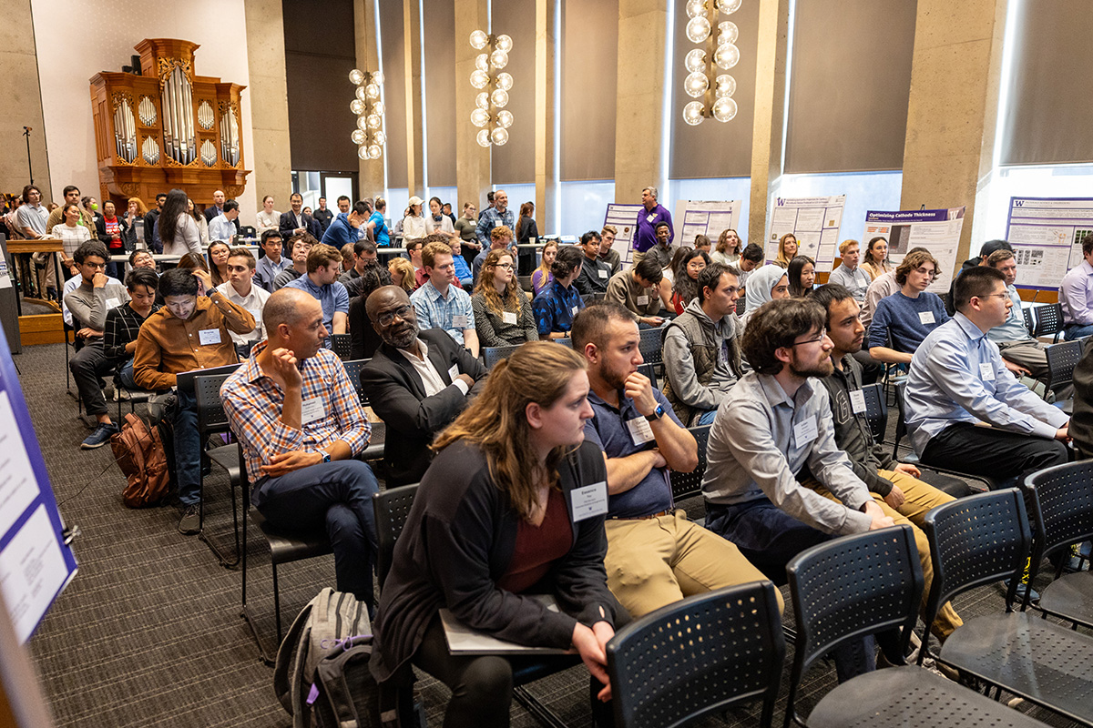 A crowd of people, some seated, some standing at the back of the room, all facing a presenter who is out of frame.