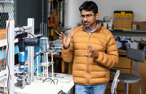 Assistant Professor Aniruddh Vashisth stands in a lab environment next to two machines used for automated welding.