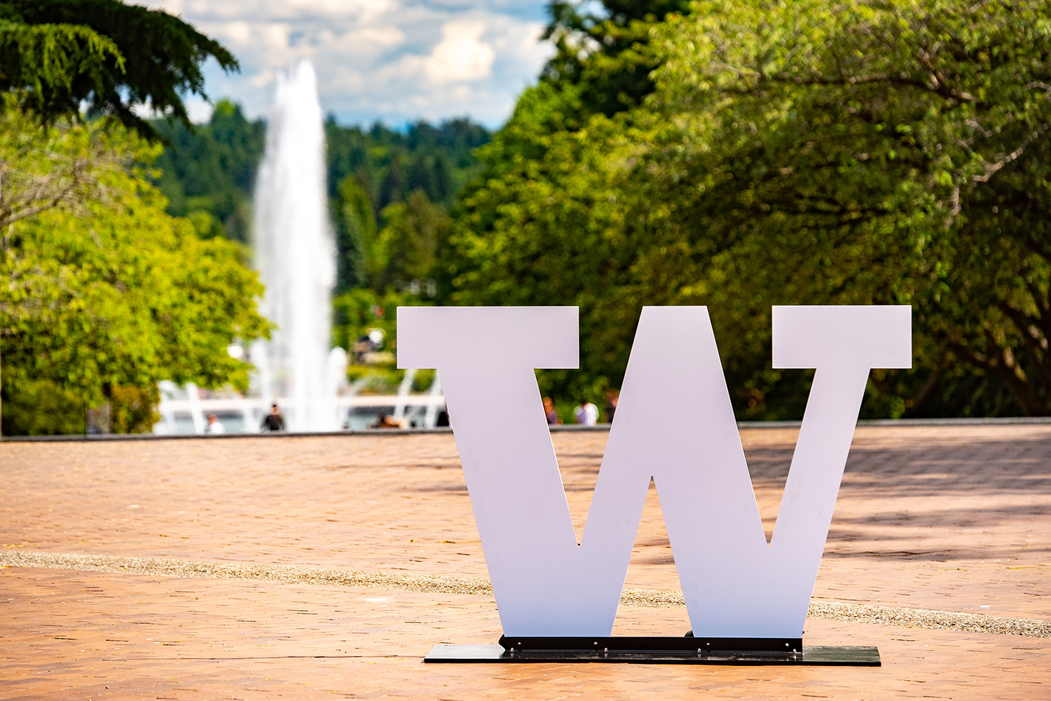 A white University of Washington 'W' logo standing on a red brick surface with Drumheller Fountain in the background.