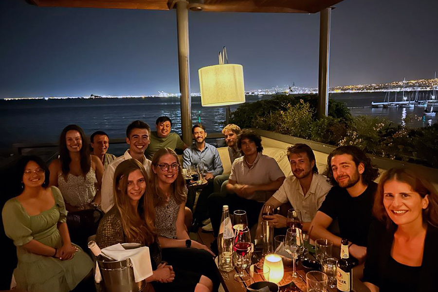 A group of students in a restaurant smile at the camera. The ocean can be seen through the windows behind them.