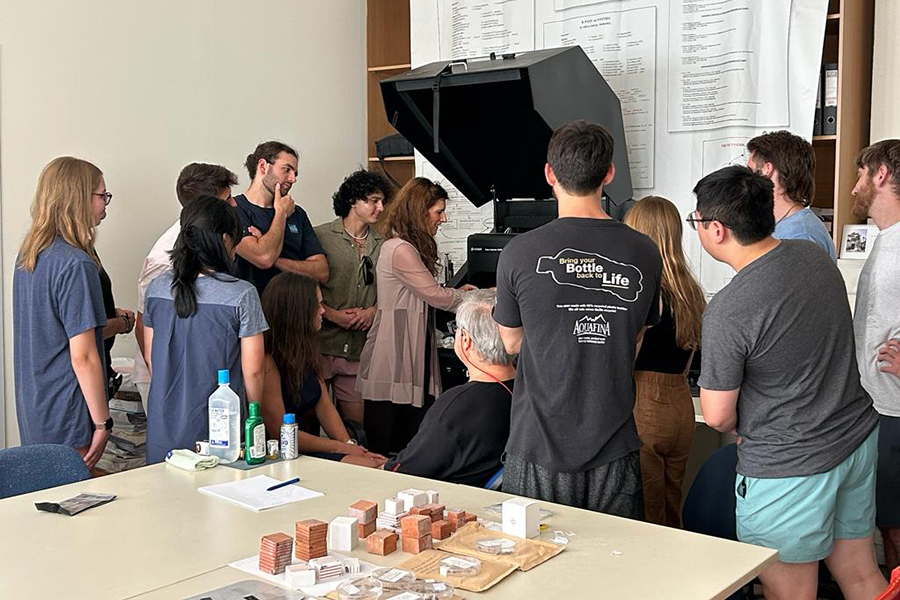 A female professor stands at a piece of equipment, demonstrating how to use it to a group of about a dozen students.