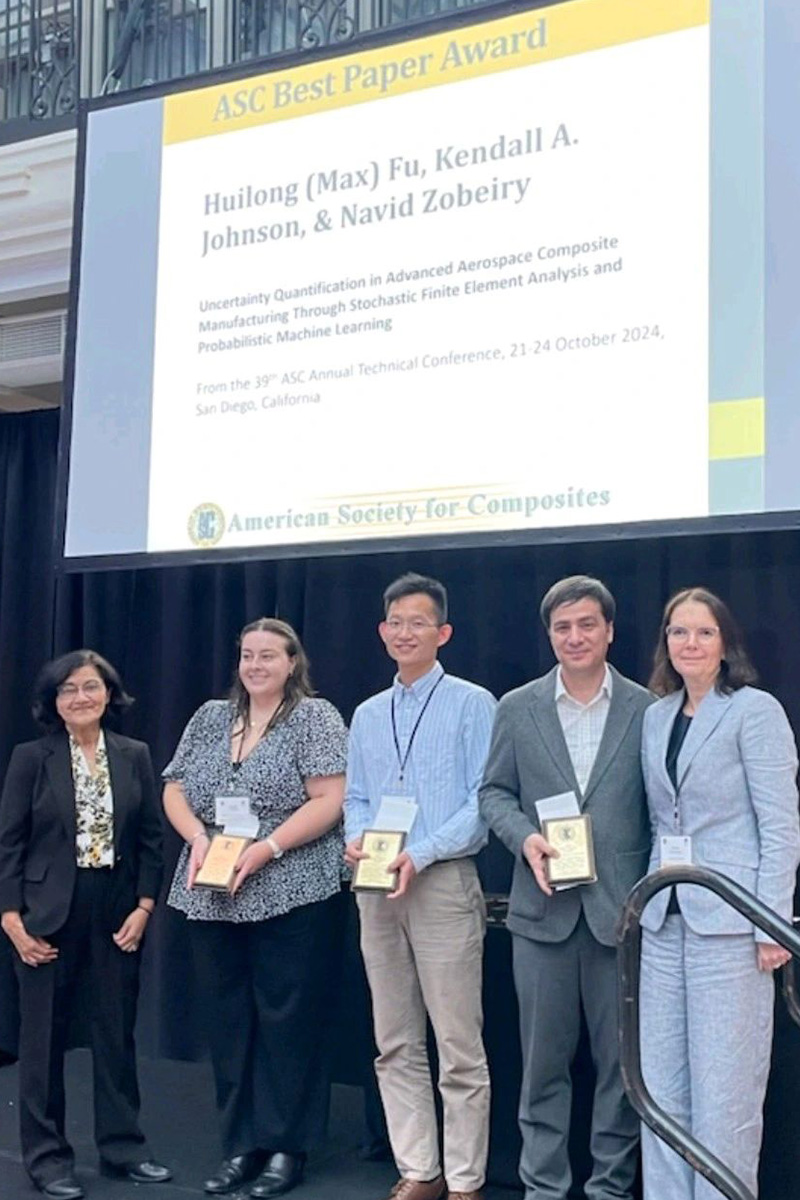 Five people in business attire standing on a stage, three holding plaques, all smiling at the camera