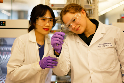 Two female students wearing white lab coats, purple gloves, and lab goggles, hold up a small lithium coin cell.