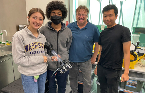 Three students and one faculty member stand in a lab setting. One student holds an arm attachment for a robot.